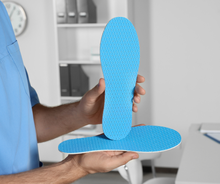 Male doctor on the left side in a doctors office holding up 2 customs blue orthotics